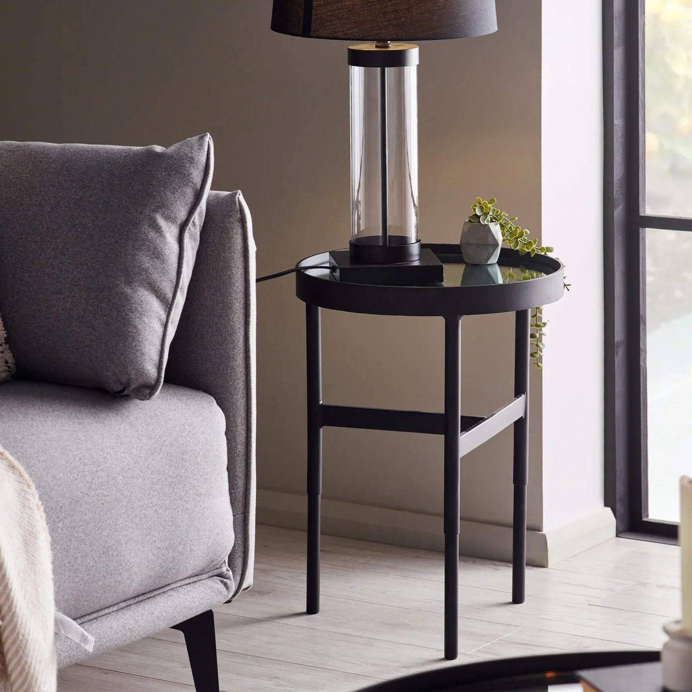 Glass lamp on black metal mirror side table next to grey sofa in bright living room