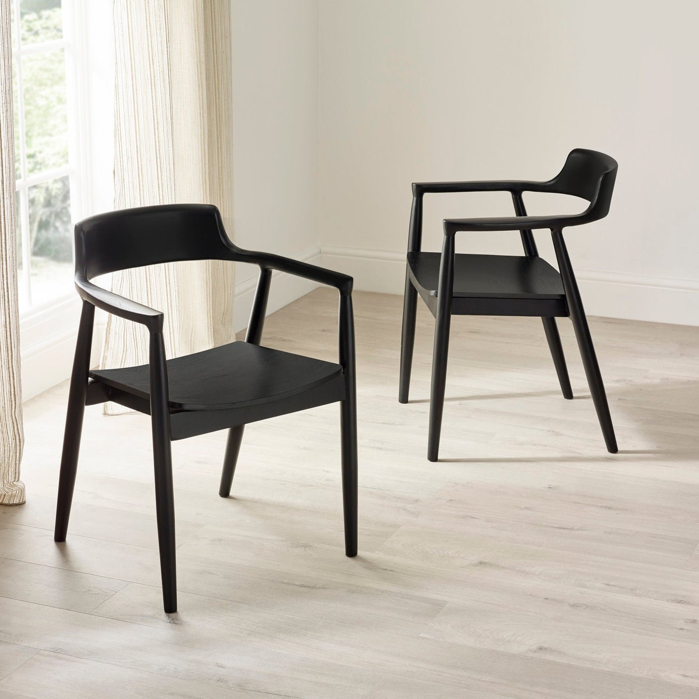 Two black wooden dining chairs with curved backs and armrests on pale wooden floor near cream curtains