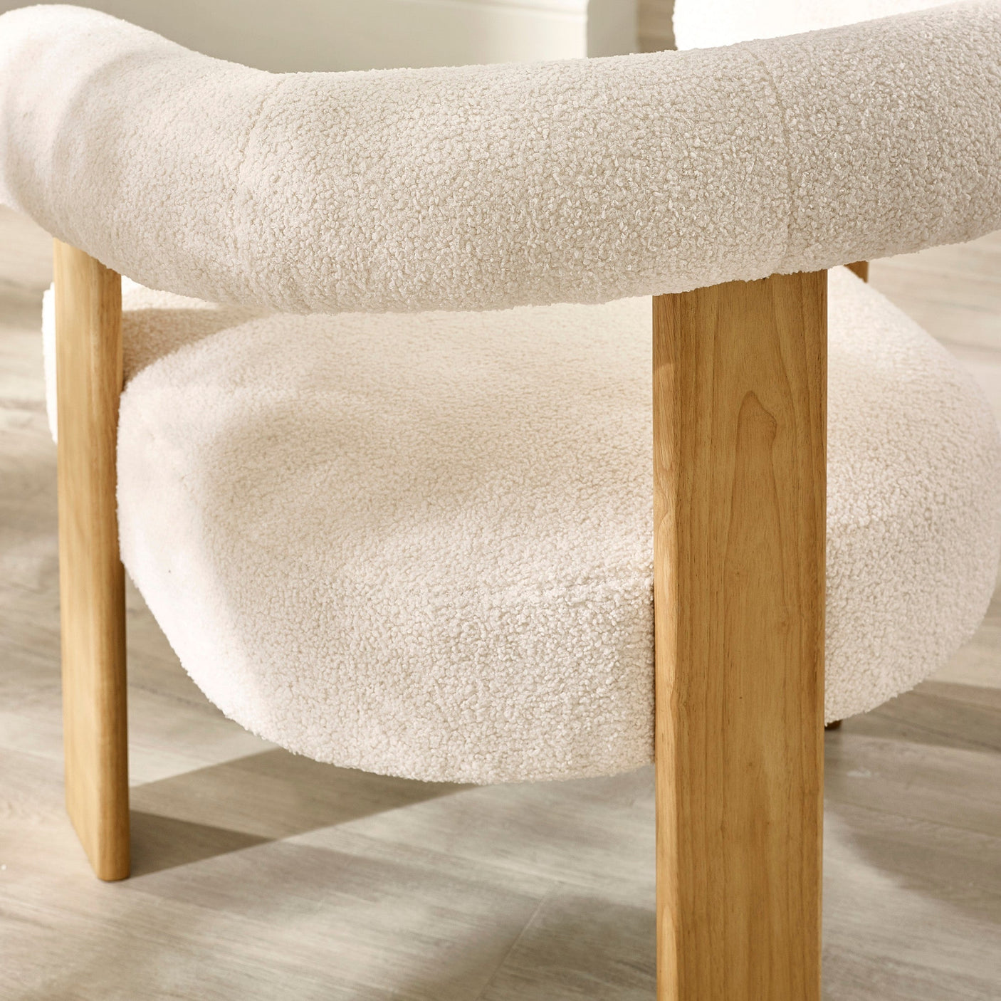 Close-up of curved boucle fabric backrest and thick oak legs of cream accent armchair on wood flooring.
