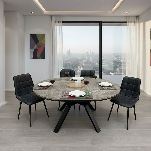 A modern round dining table with a gray stone-look top and black angled legs, surrounded by four black upholstered dining chairs in a bright contemporary dining room with large windows overlooking a city skyline.