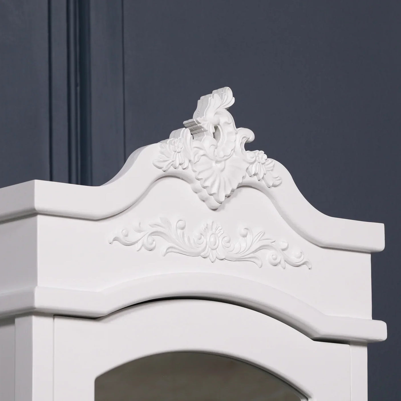 White ornate wardrobe top with decorative floral carvings against dark wall