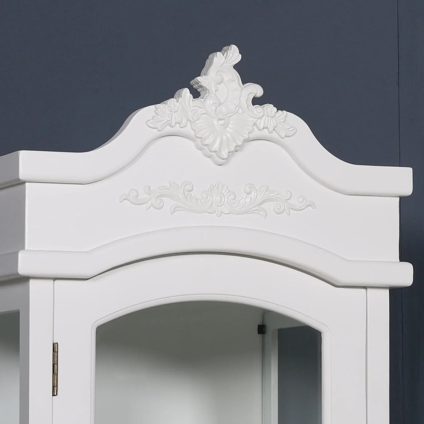 White display cabinet with ornate carved detailing and glass door against blue wall