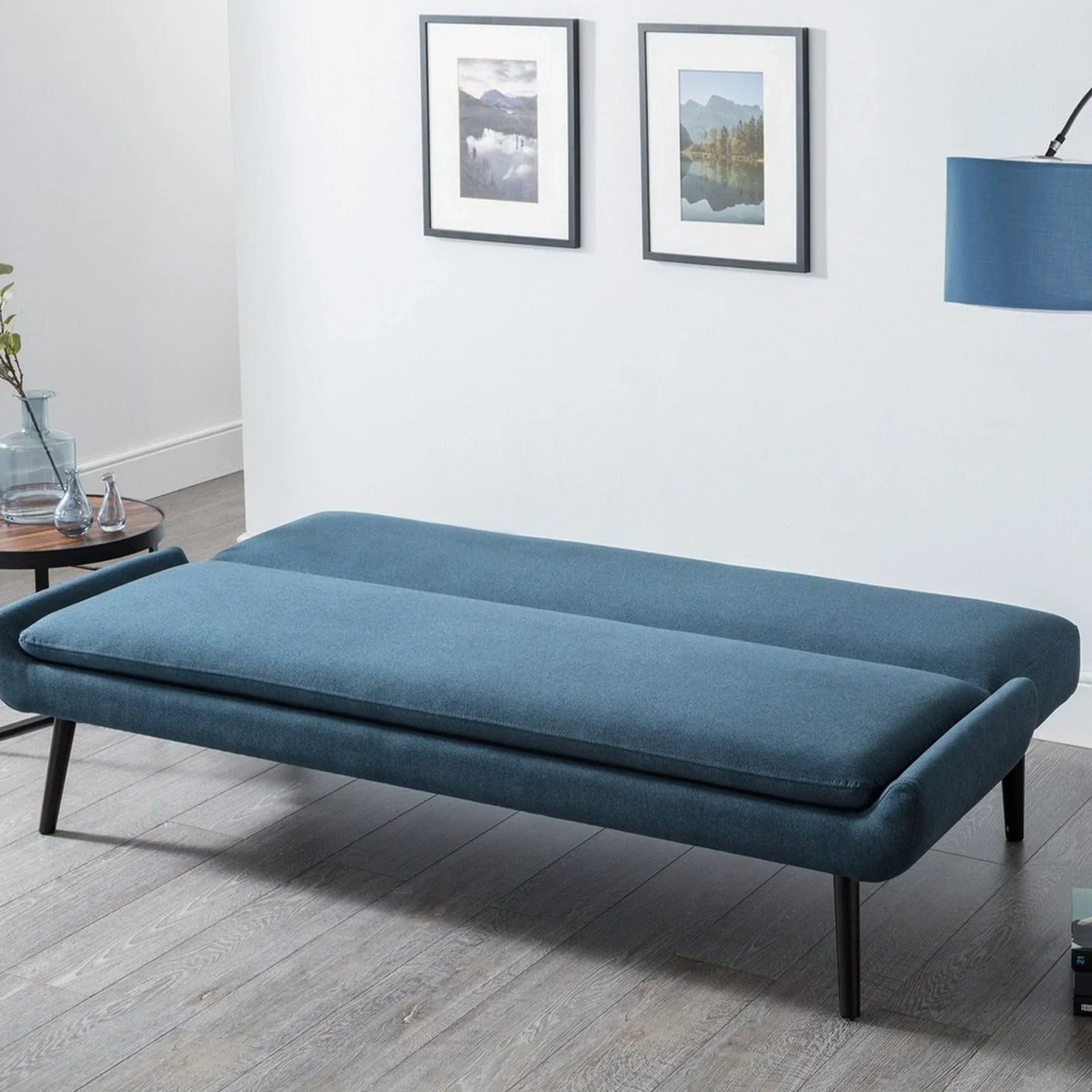 Blue sofabed with black legs in modern room with wood floor and two framed wall prints