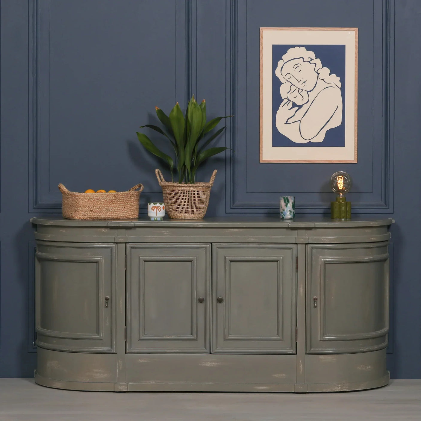 Grey sideboard cabinet with paneled doors styled with baskets, plant, and lamp against blue wall
