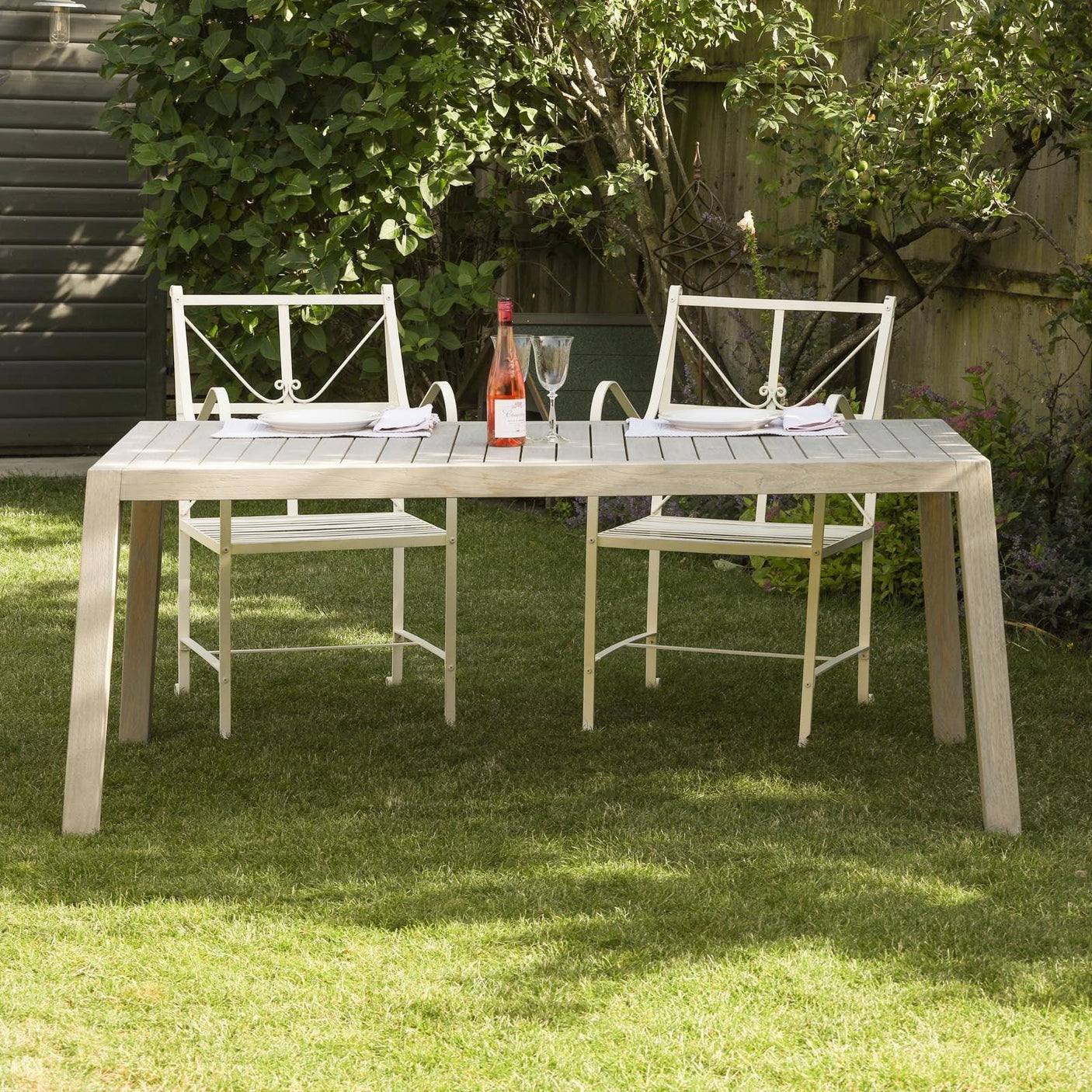 Wooden garden dining table with two white metal chairs set on lawn