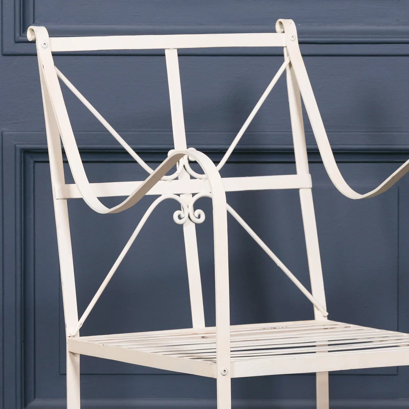 Cream metal garden chair with slatted seat and decorative scroll backrest against blue wall