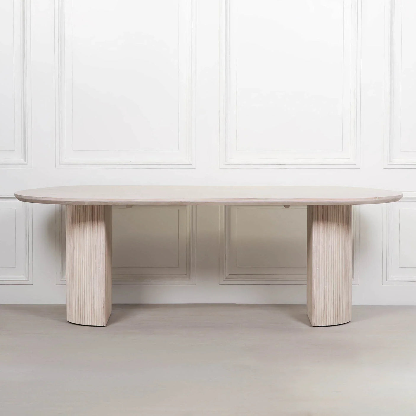 Light wood dining table with oval top and two fluted pedestal legs against white wall