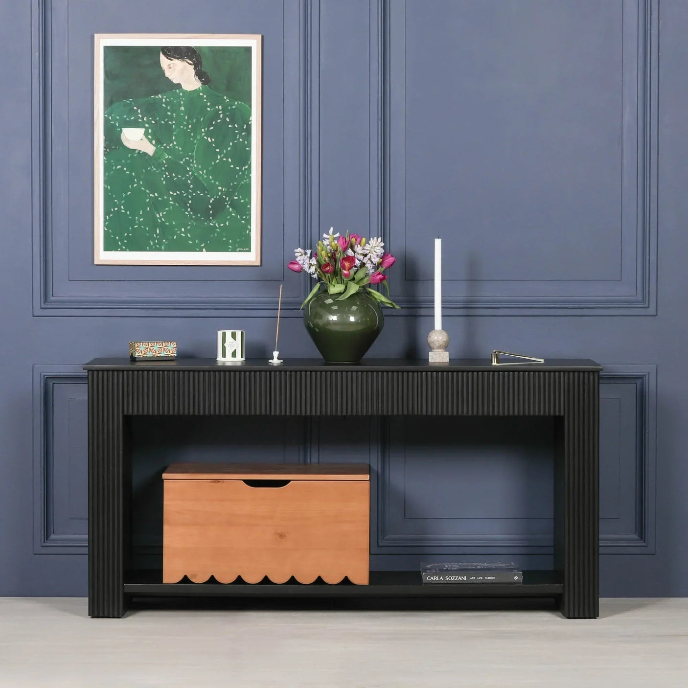 Black console table with fluted design against blue wall, wood storage box underneath, decor on top