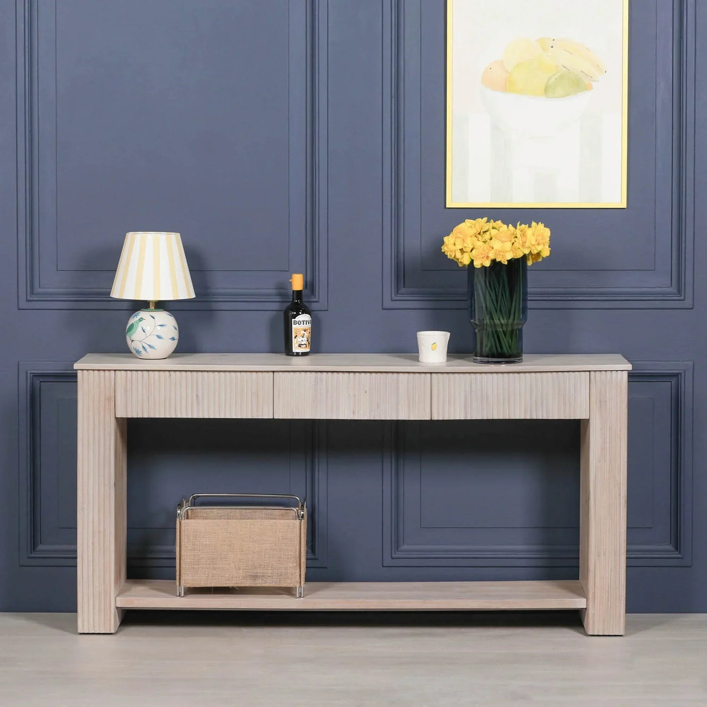 Light wood console table with drawer, vase, lamp, and magazine rack against blue paneled wall