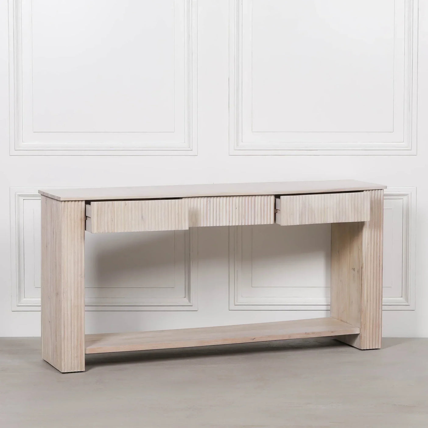 Light wood console table with grooved drawer fronts and lower shelf against white panel wall