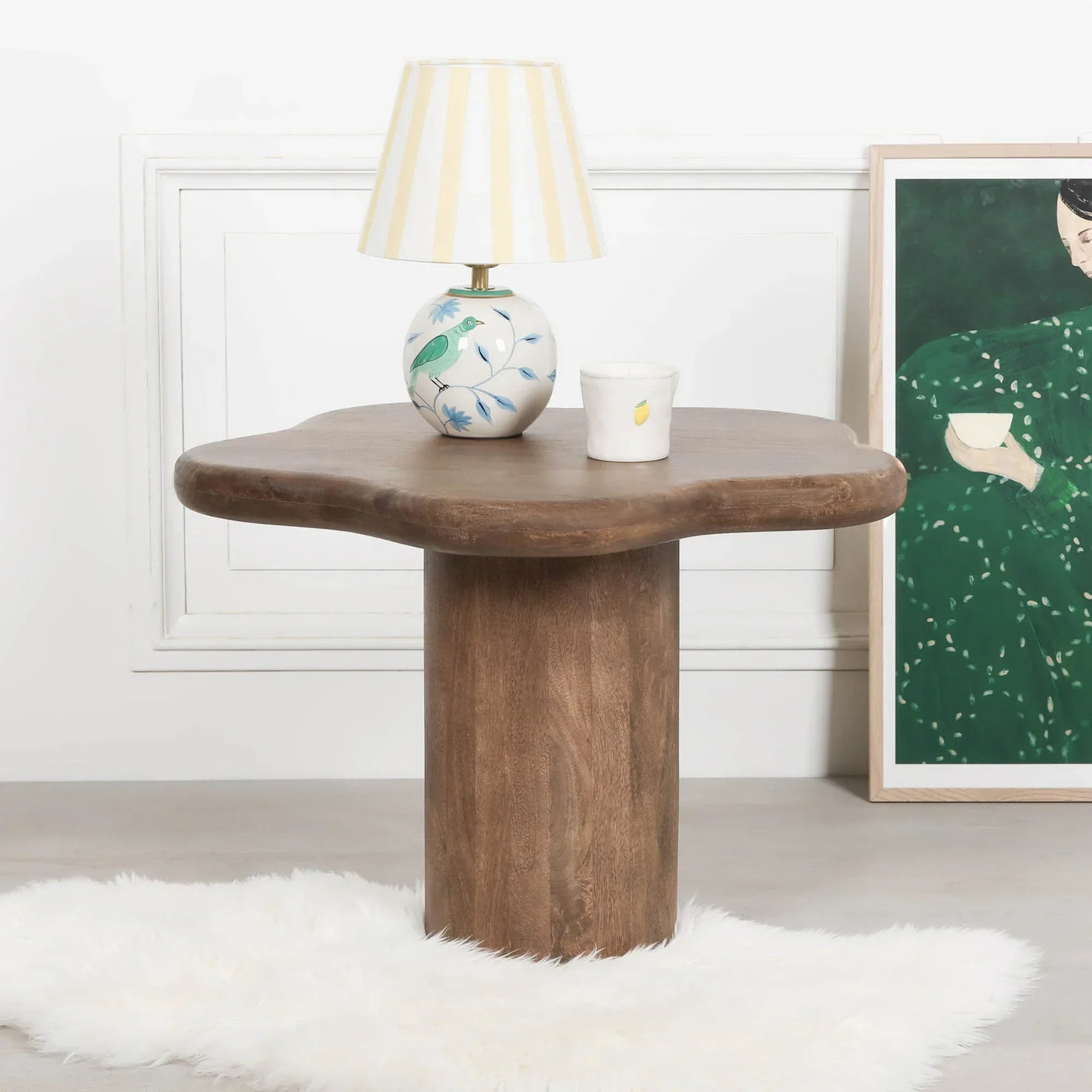 Brown wooden side table with sculpted edge, lamp and cup on top, white faux fur rug