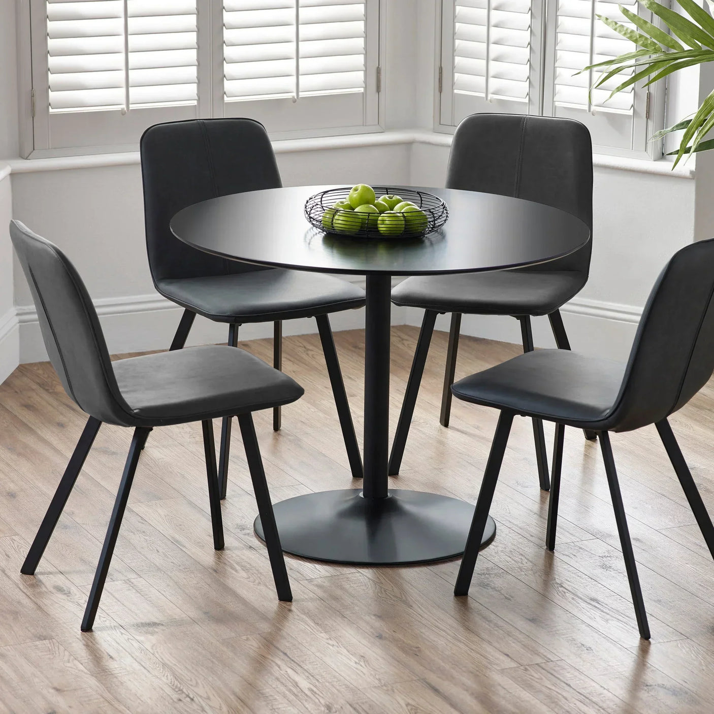 Round black dining table with four black chairs on wood floor, bowl of green apples on top