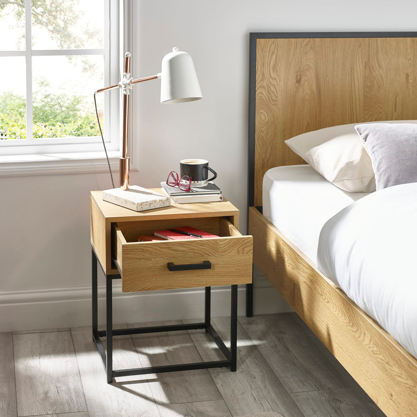 Oak bedside table with black metal legs, open drawer, lamp and accessories beside a bed