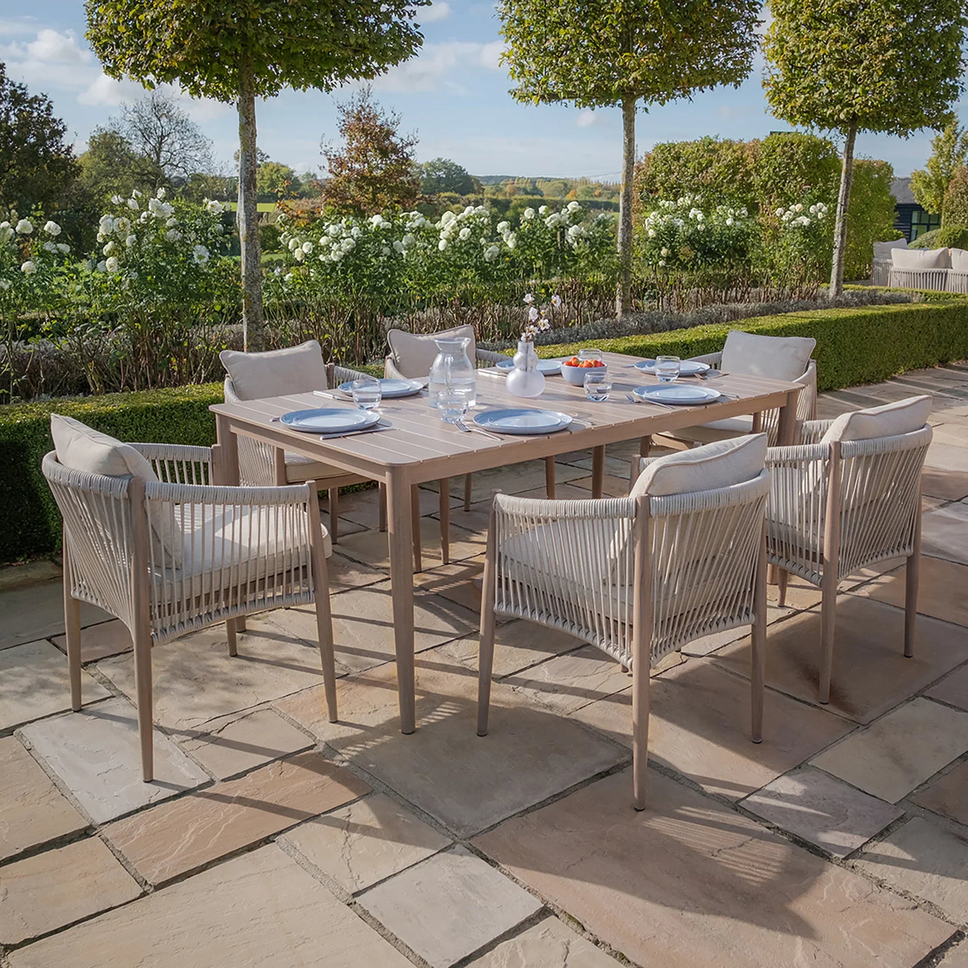 Outdoor dining set with six cushioned rope chairs and wooden table on stone patio