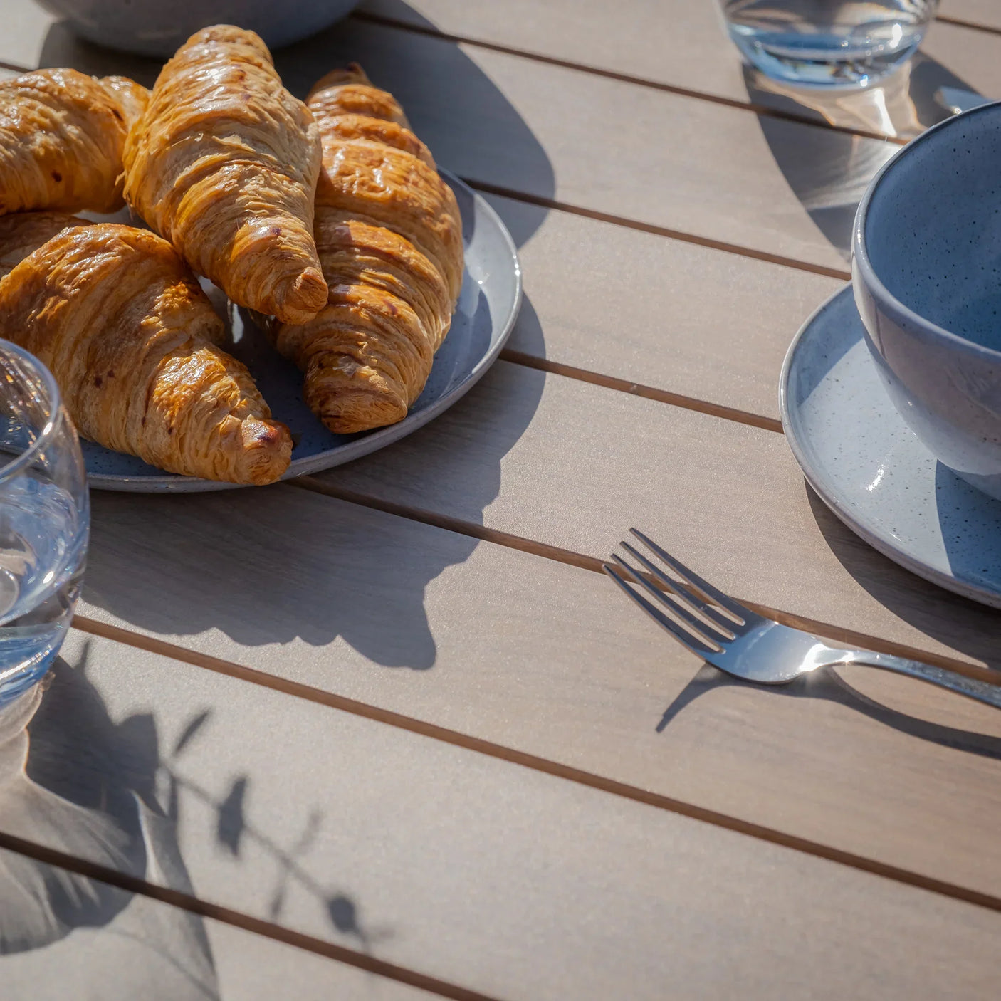 Light wood outdoor dining table with grey plates, bowls, croissants and glass of water