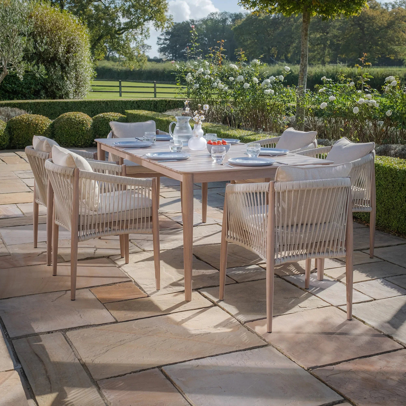 Wooden outdoor dining table set with six rope armchairs and cream cushions on patio