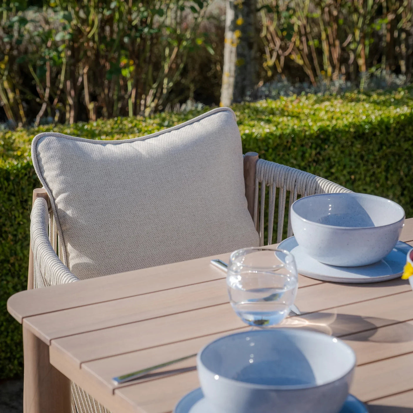 Beige rope outdoor dining chair with cushion beside light wood garden table and tableware