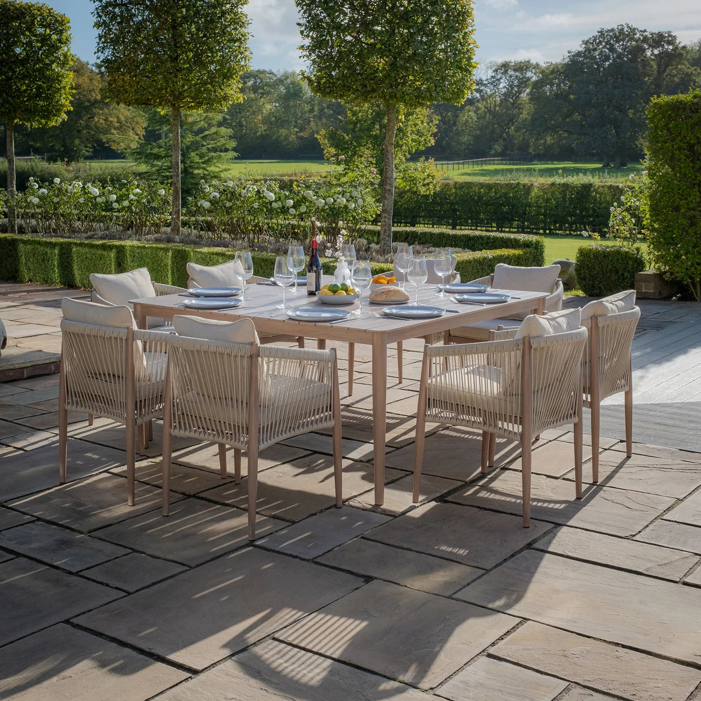 Wooden outdoor dining set with eight rope-back chairs on stone patio, table set for meal