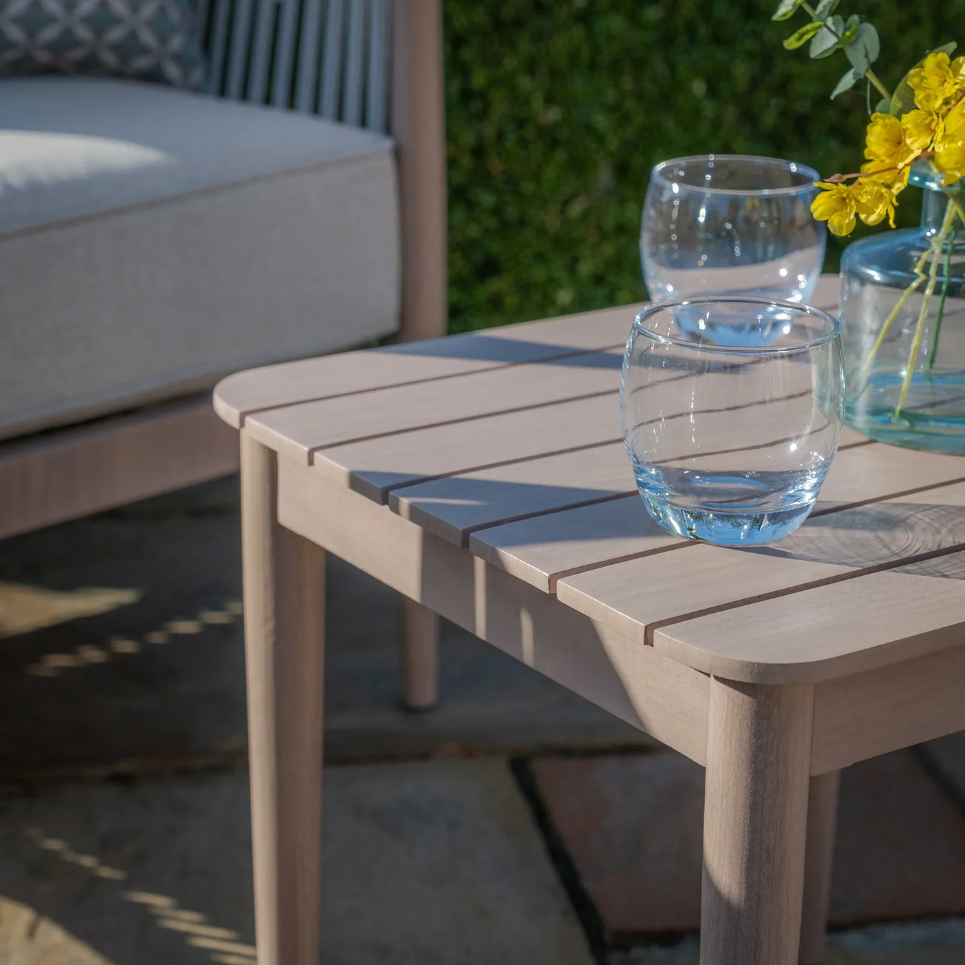 Light wood outdoor coffee table with slatted top, glassware and vase with yellow flowers