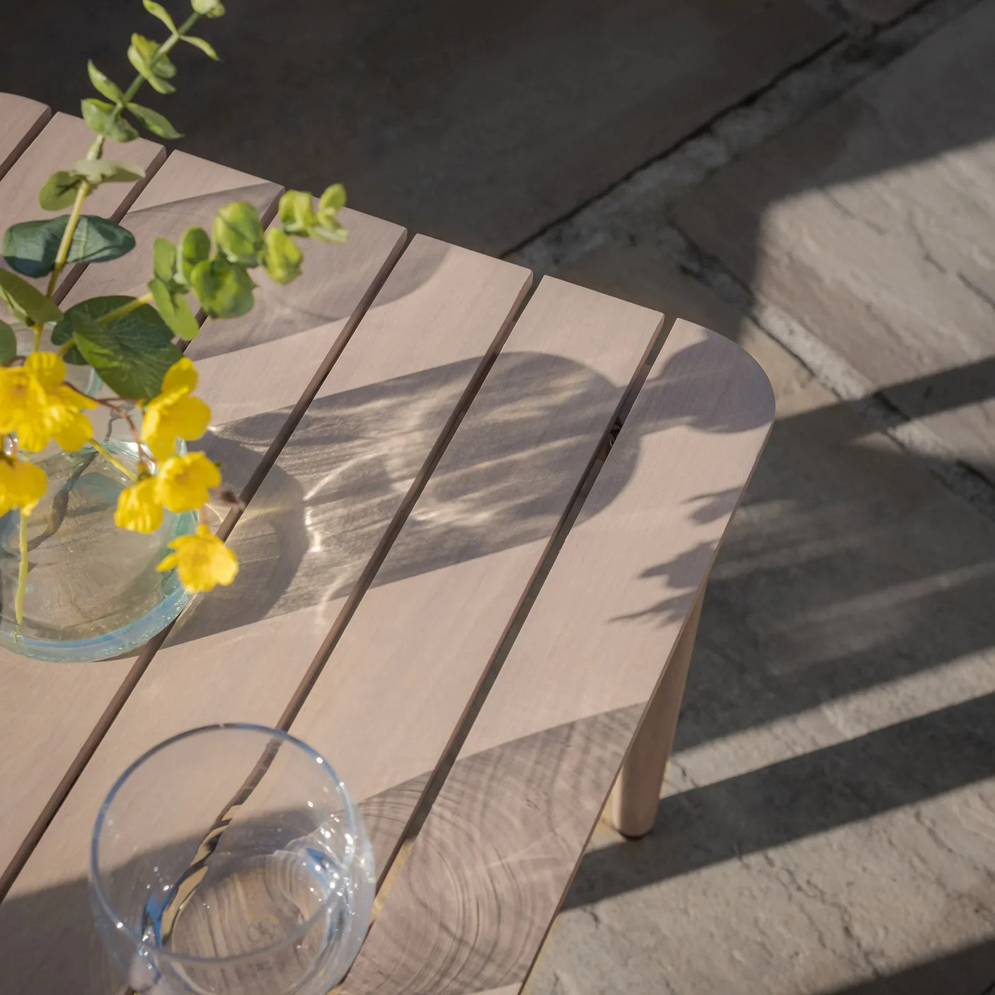 Light wood slatted outdoor coffee table with glass vase of yellow flowers and clear drinking glass