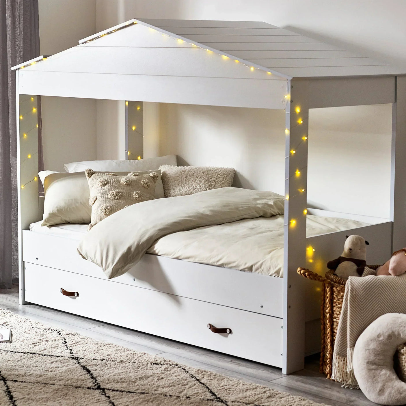 White low sleeper bed frame with house-shaped canopy, trundle drawer, fairy lights, and bedding