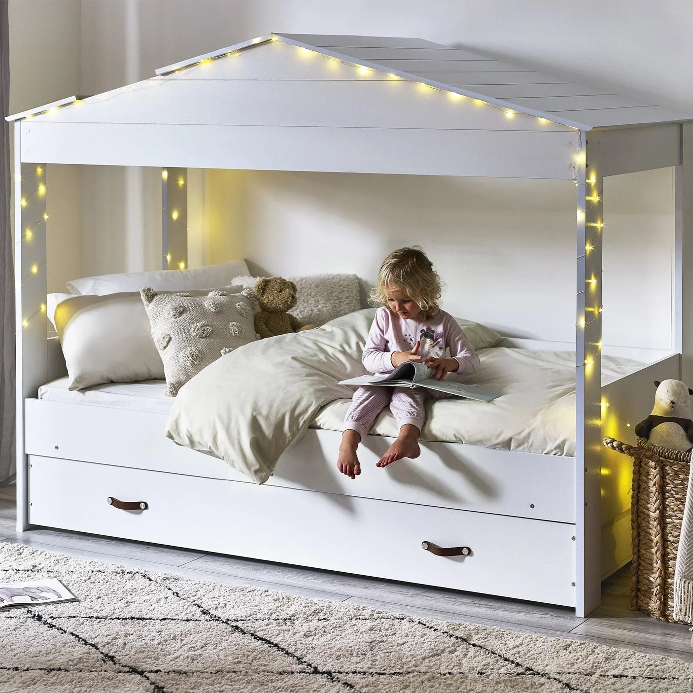 White low sleeper bed with trundle drawer, canopy frame, fairy lights, and child reading
