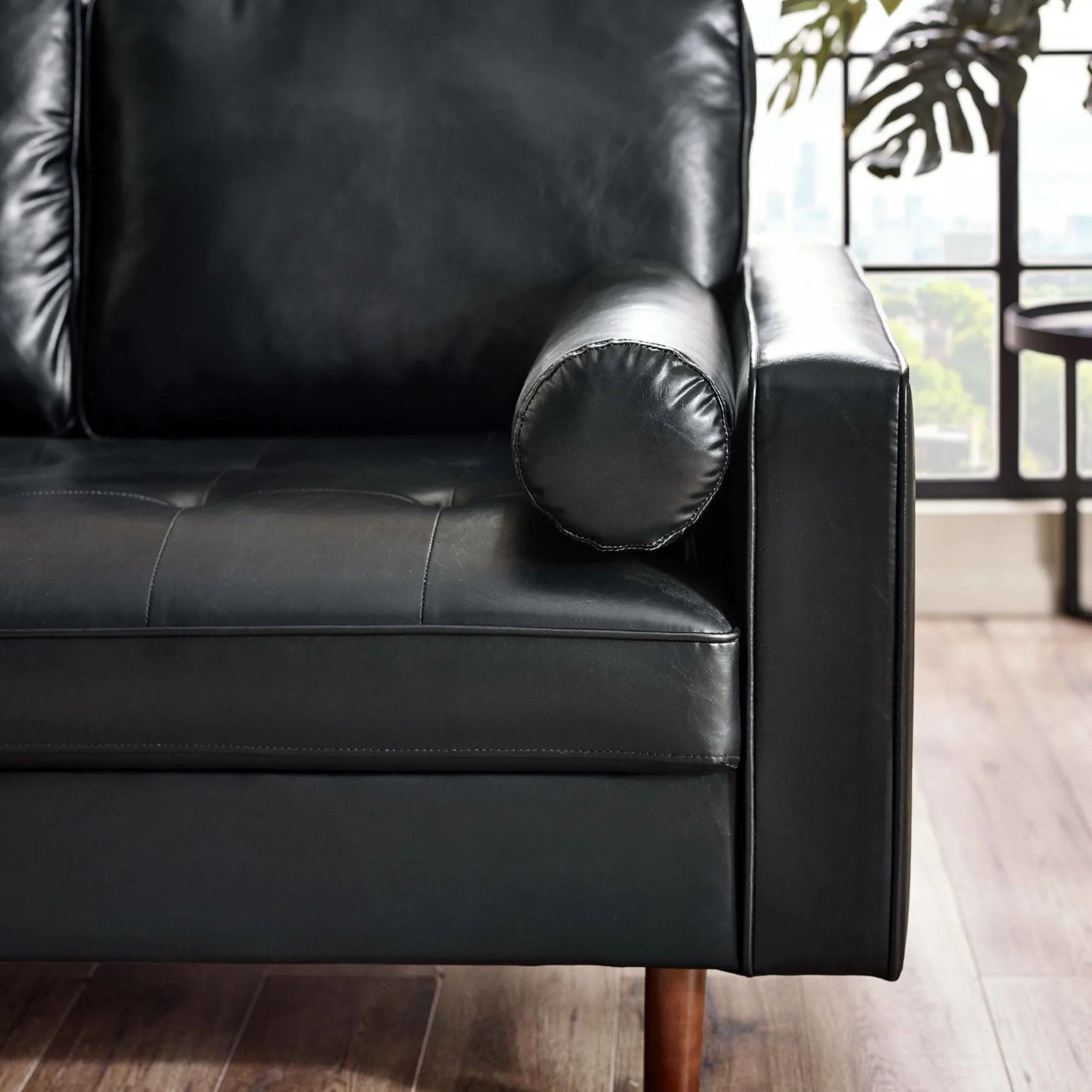 Black leather sofa closeup with round bolster armrest and wooden legs near window