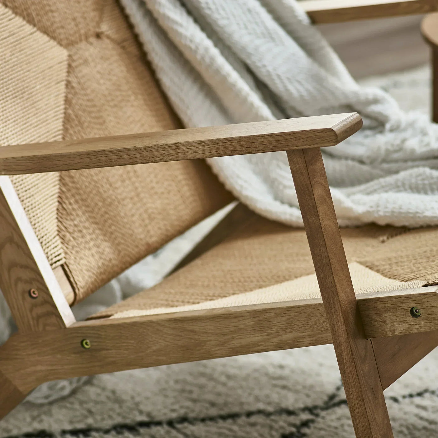 Close up of woven lounge chair with wooden armrest and beige throw on textured rug