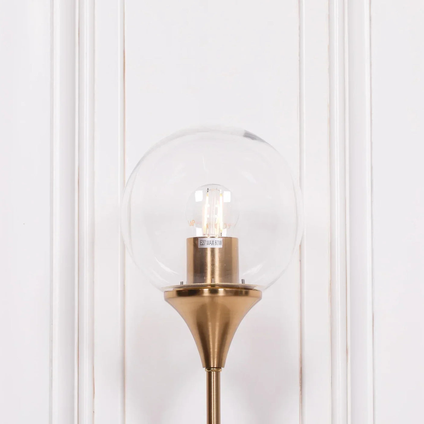 Brass wall light with clear glass globe shade against white panel wall