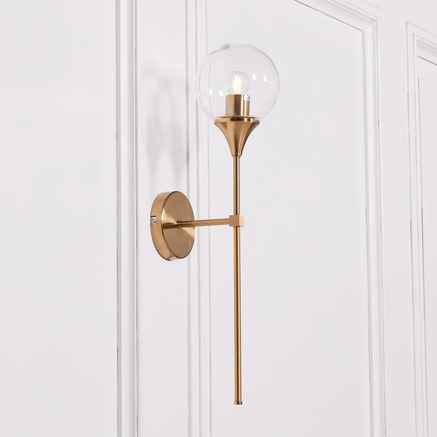 Gold wall light with clear glass globe shade mounted on white panelled wall