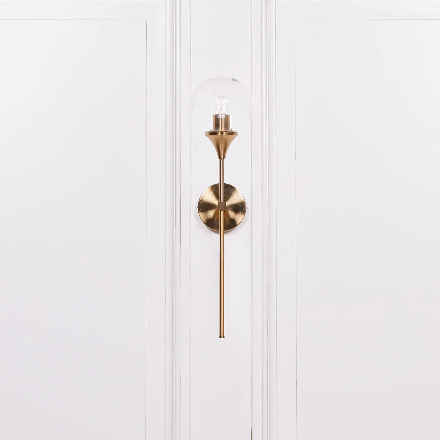 Brass wall sconce with clear glass shade mounted on white panelled wall