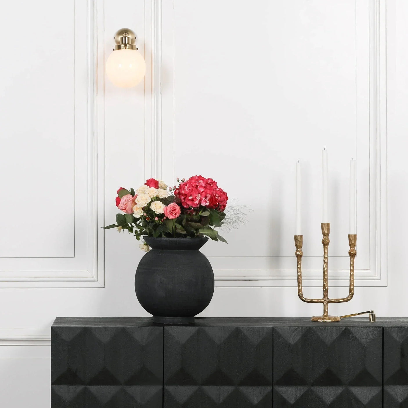 Modern wall light with globe shade above black sideboard, flowers and gold candle holder