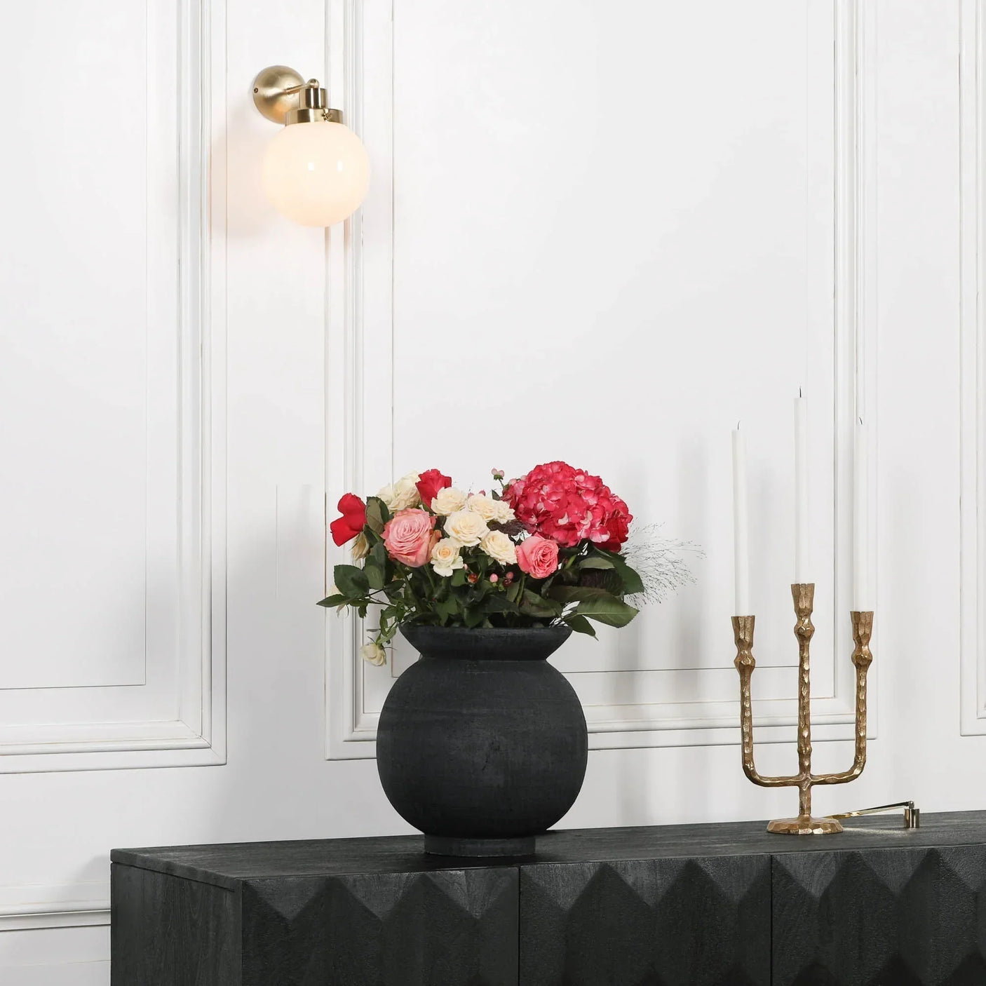 Black vase with pink and cream flowers on dark sideboard beside gold candle holder