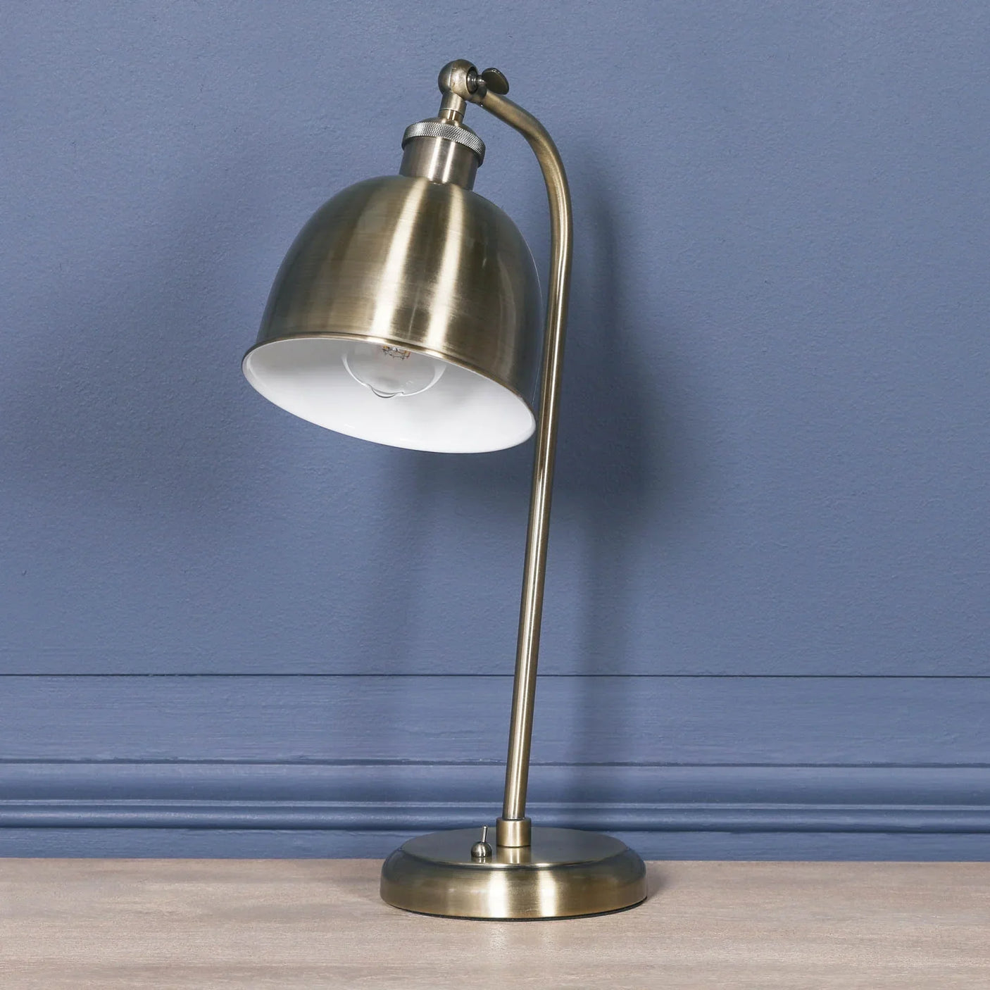 Brushed brass table lamp with curved stem and dome shade on wooden surface against blue wall