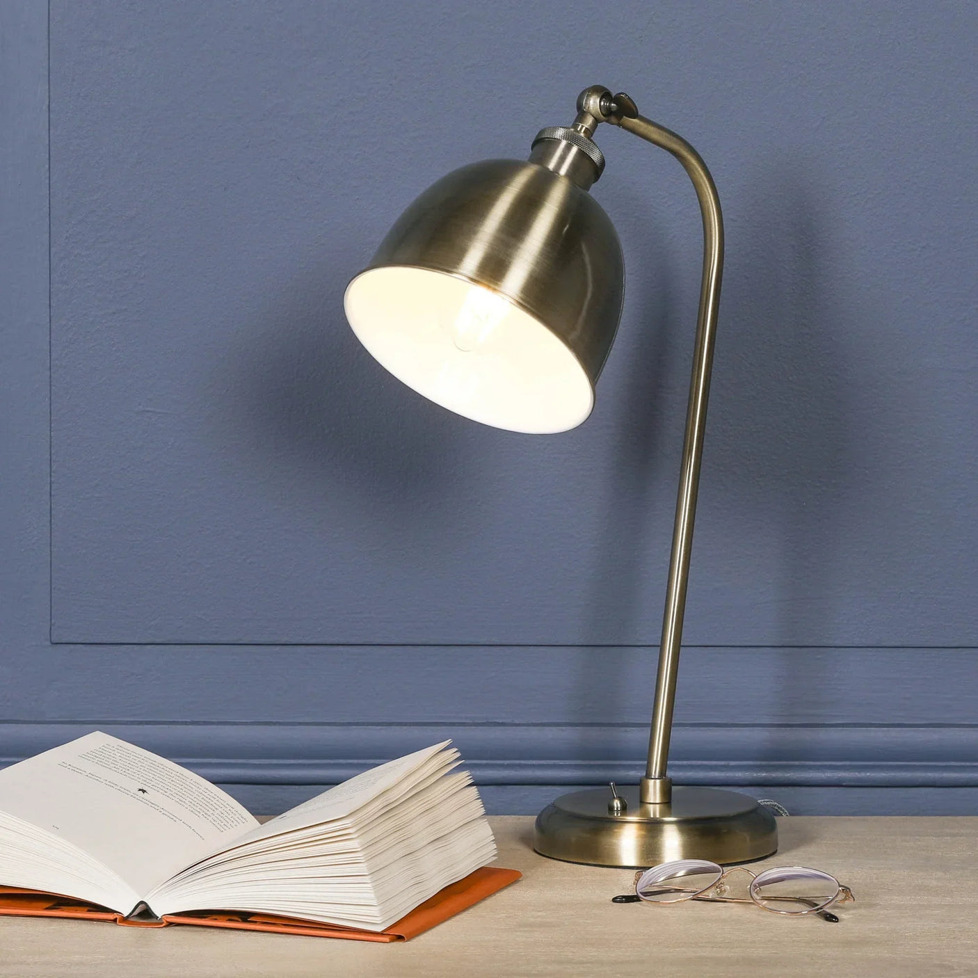 Brass desk lamp with angled shade beside open book and eyeglasses on wooden table
