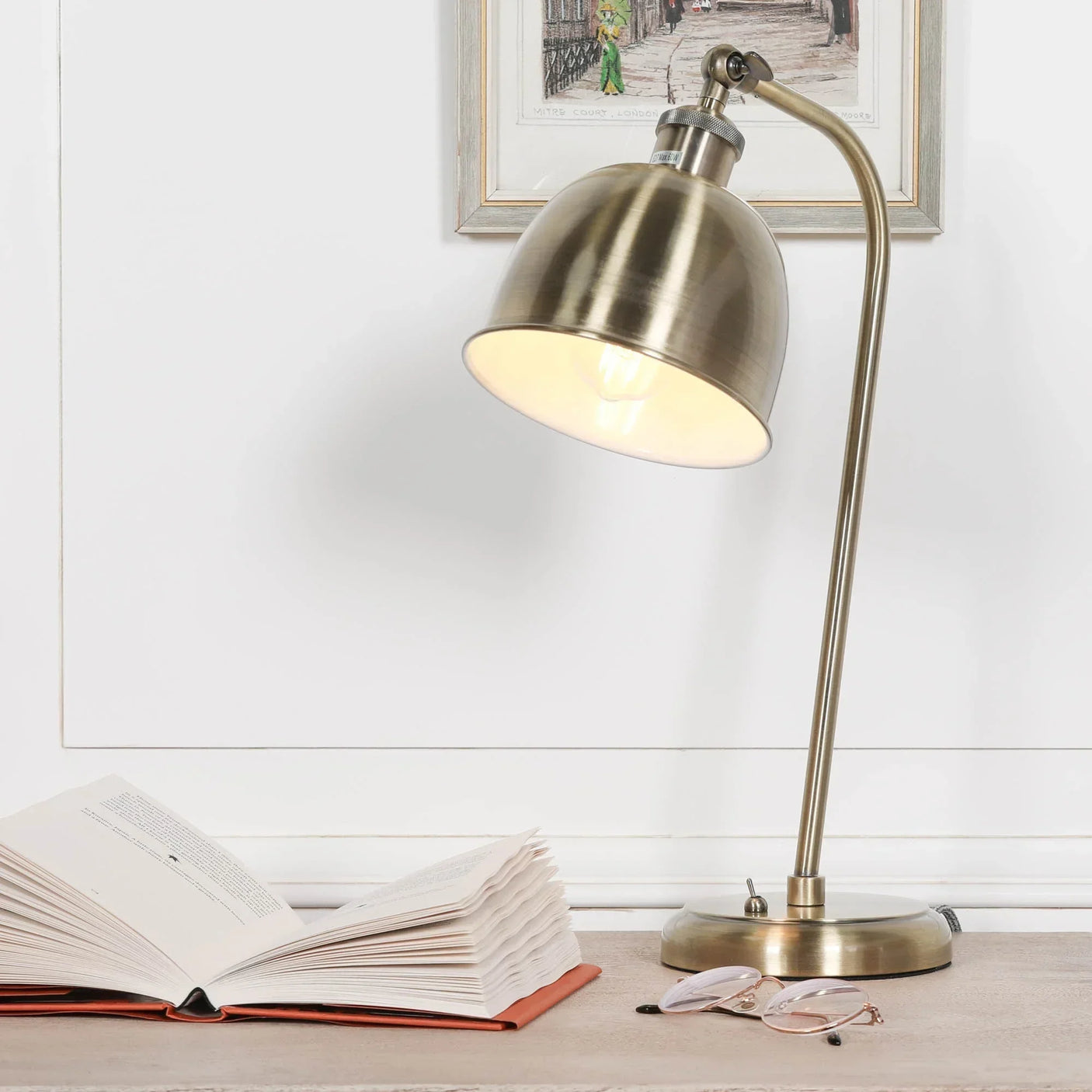 Brushed brass desk lamp with curved arm beside open book and eyeglasses on table