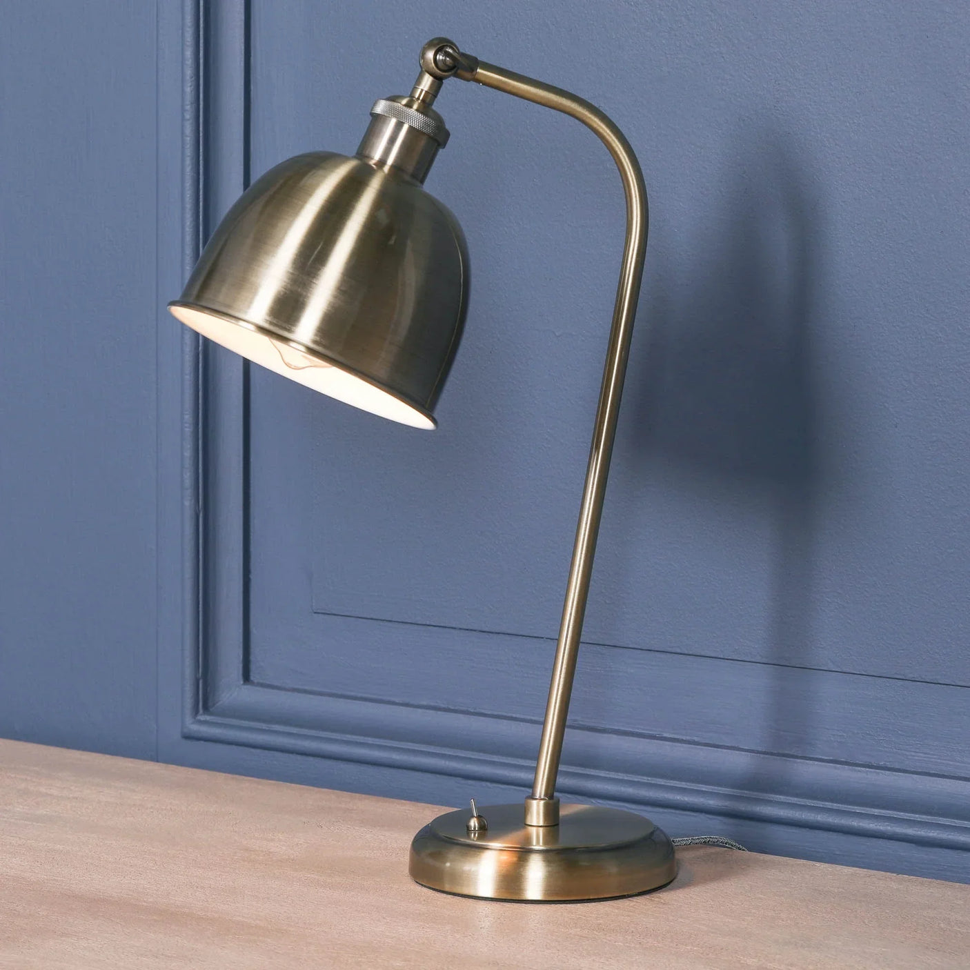 Brass finish desk lamp with adjustable dome shade on wooden table against blue wall