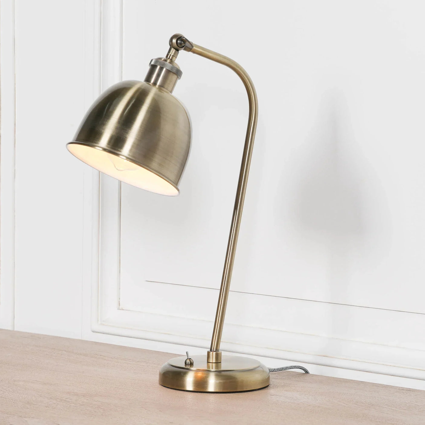 Brass desk lamp with domed shade on wooden table against white paneled wall