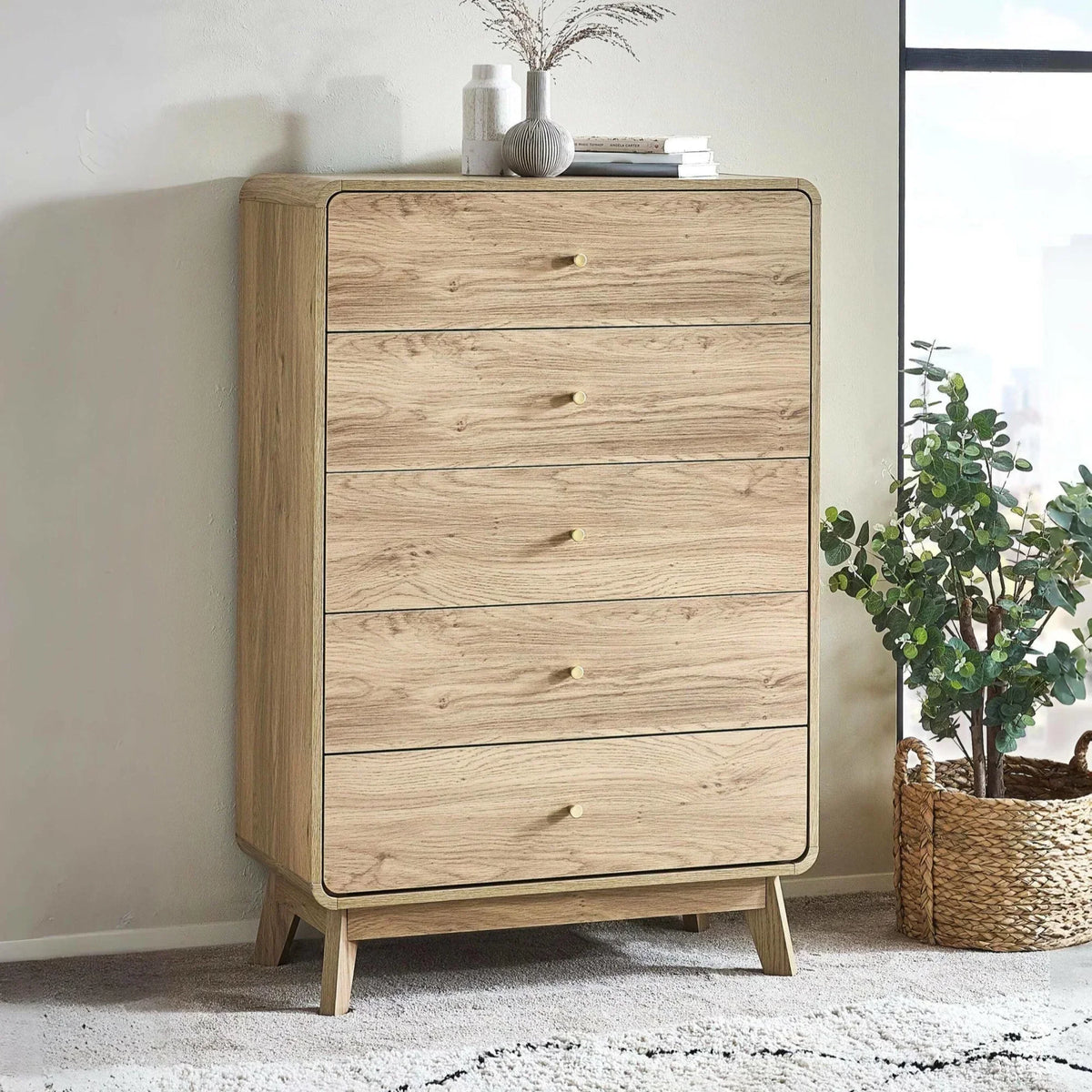 Light oak chest of drawers with five drawers, brass knobs, styled next to a plant basket