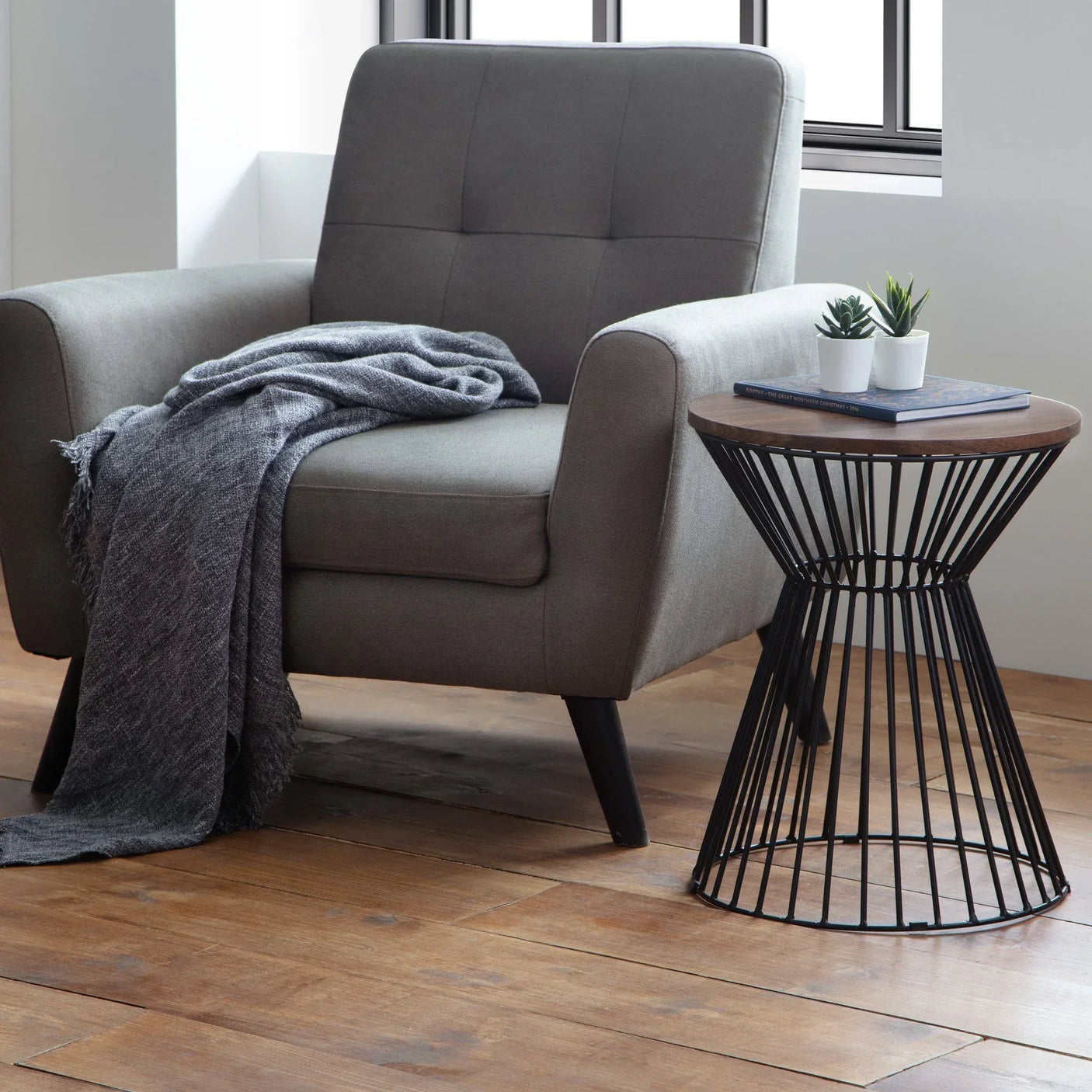 Grey accent chair with throw beside black metal wire side table with wood top and houseplants