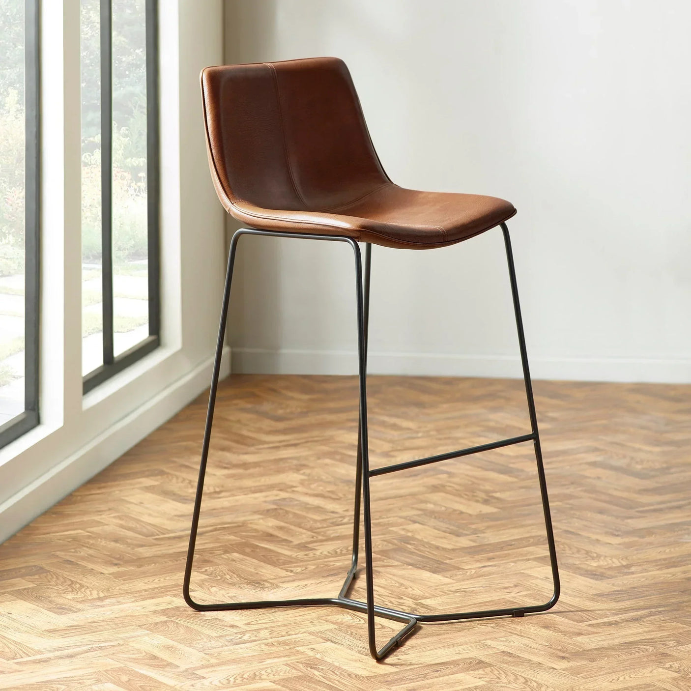 Brown leather bar stool with black metal legs beside window on herringbone wood floor