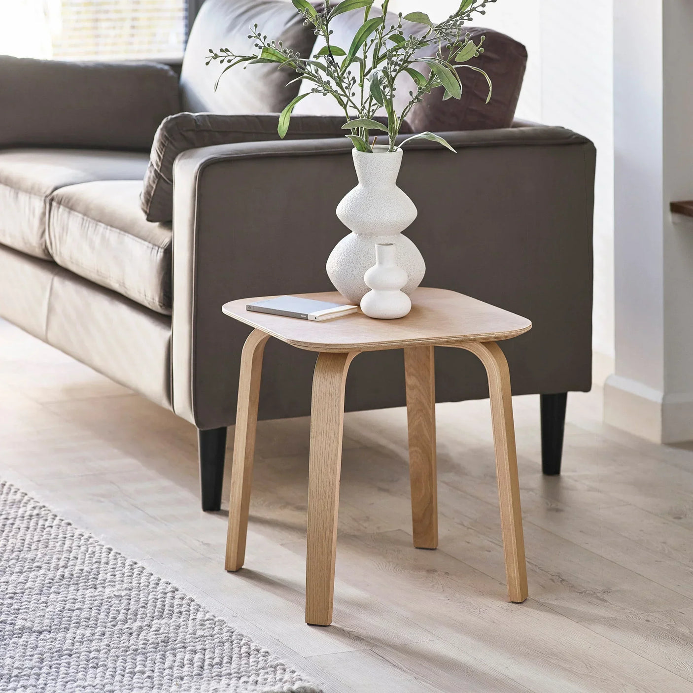 Light wood side table beside sofa, topped with white vases and greenery in modern living room