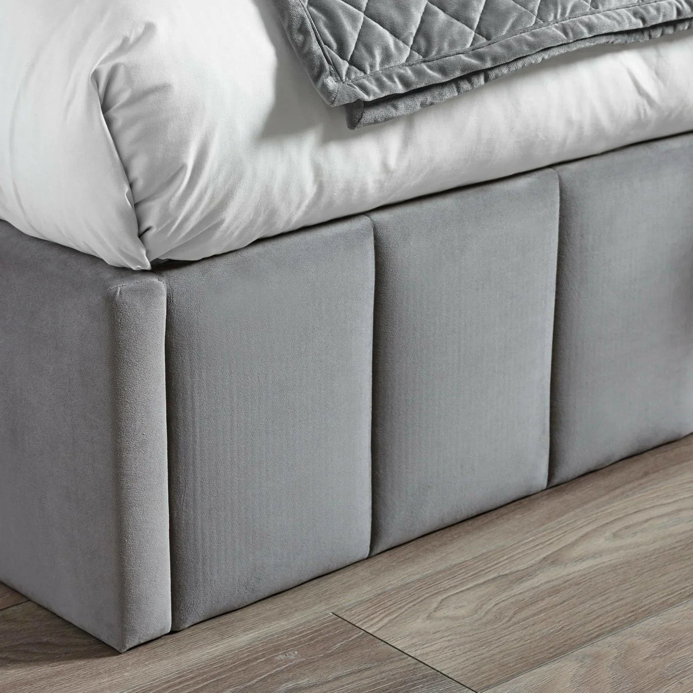 Grey velvet bed base with panel detail on wooden floor, partial bedding visible