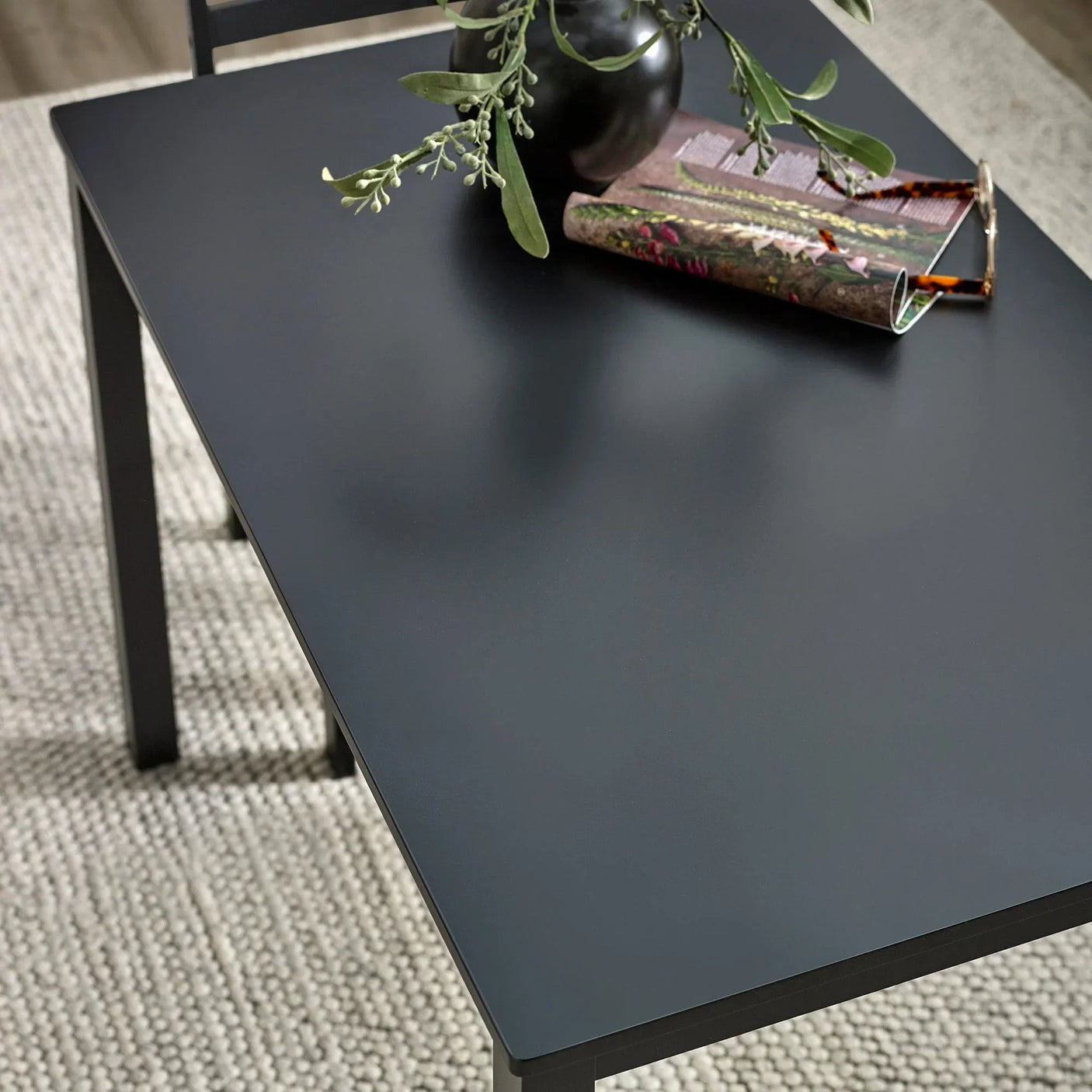Black dining table with matte finish, vase and magazine on top, shown on woven rug