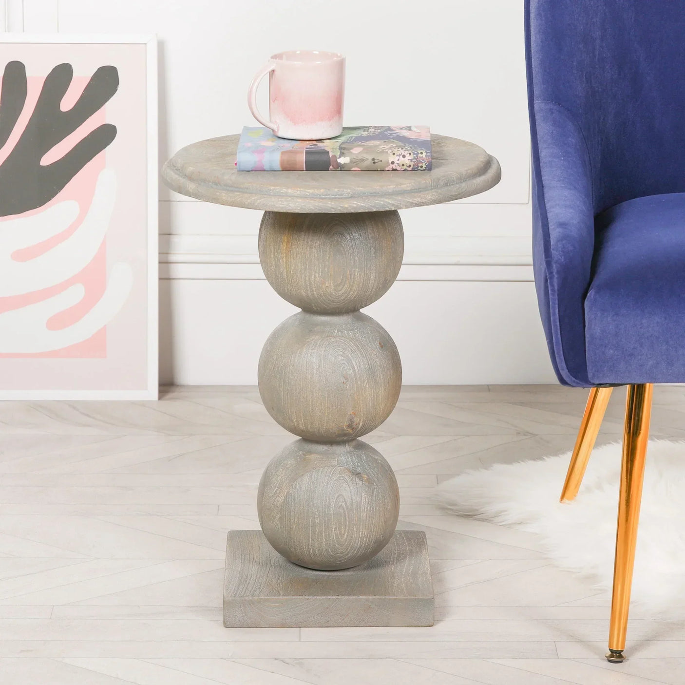 Grey wooden side table with stacked sphere base beside a blue chair and framed art print