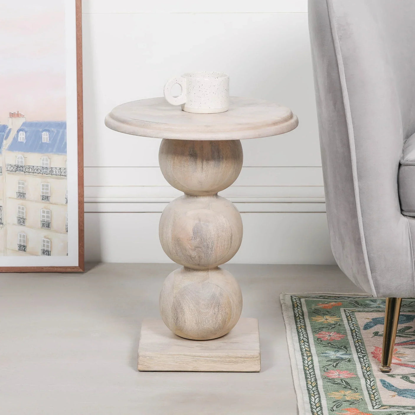 Light wood side table with sculpted round base beside sofa and rug, ceramic mug on top