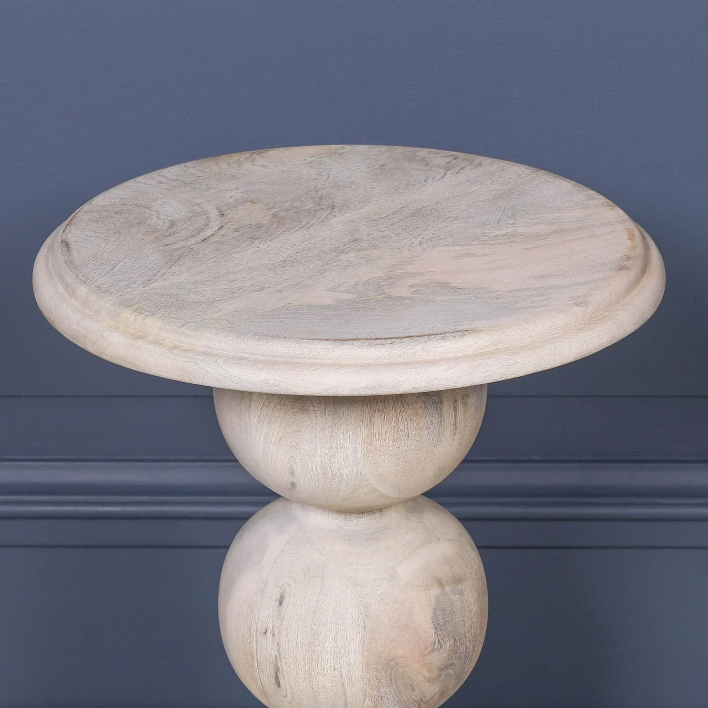 Round wooden side table with natural finish and sculpted pedestal base against blue wall