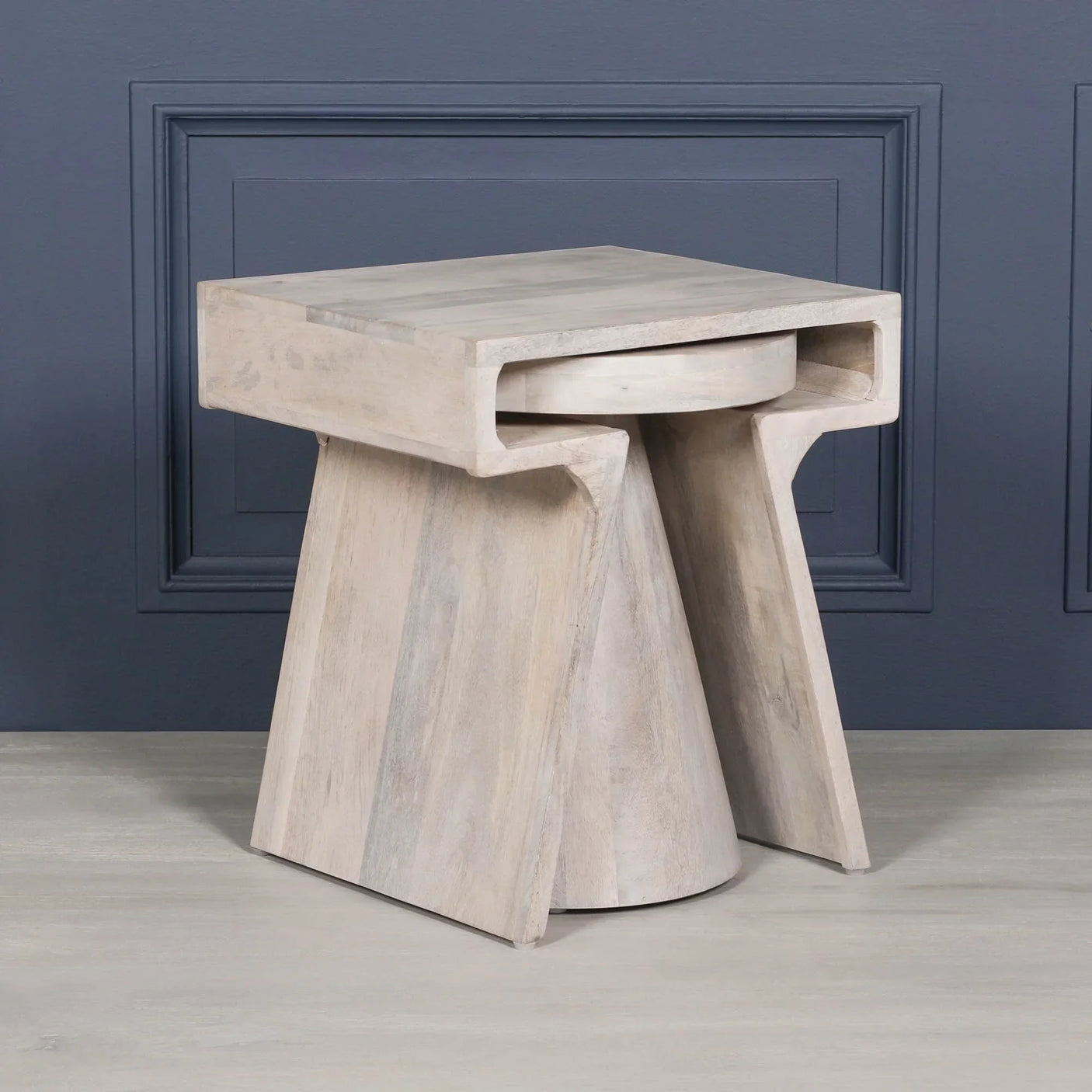 Modern light wood side table with geometric base and open shelf against navy panel wall