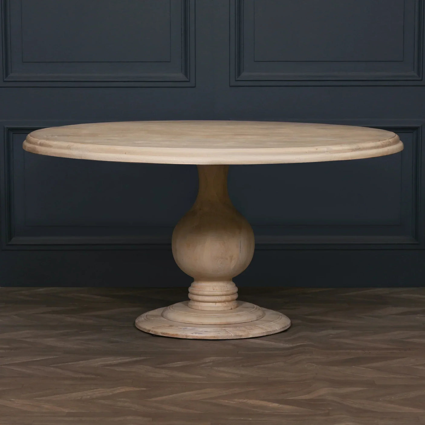 Round wooden dining table with pedestal base on herringbone floor against dark panel wall