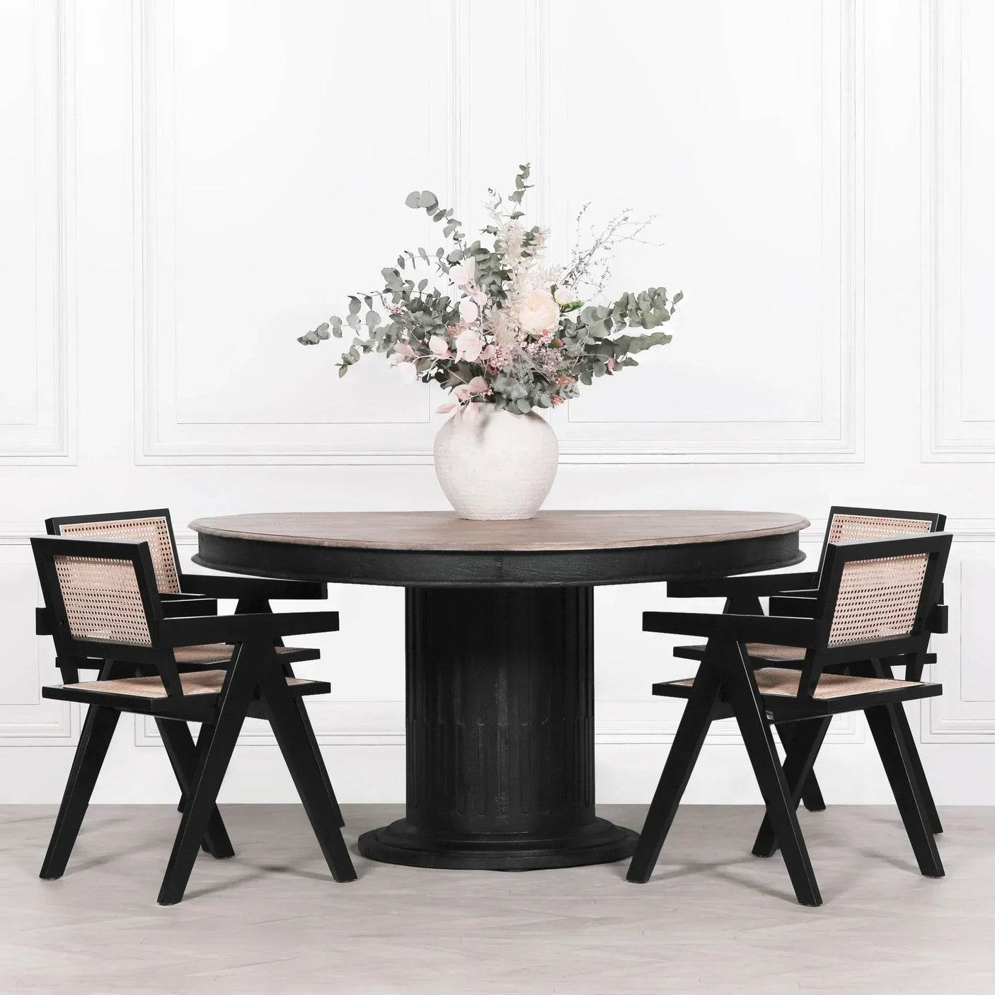 Round wood dining table with black pedestal base and four cane dining chairs, floral vase on top